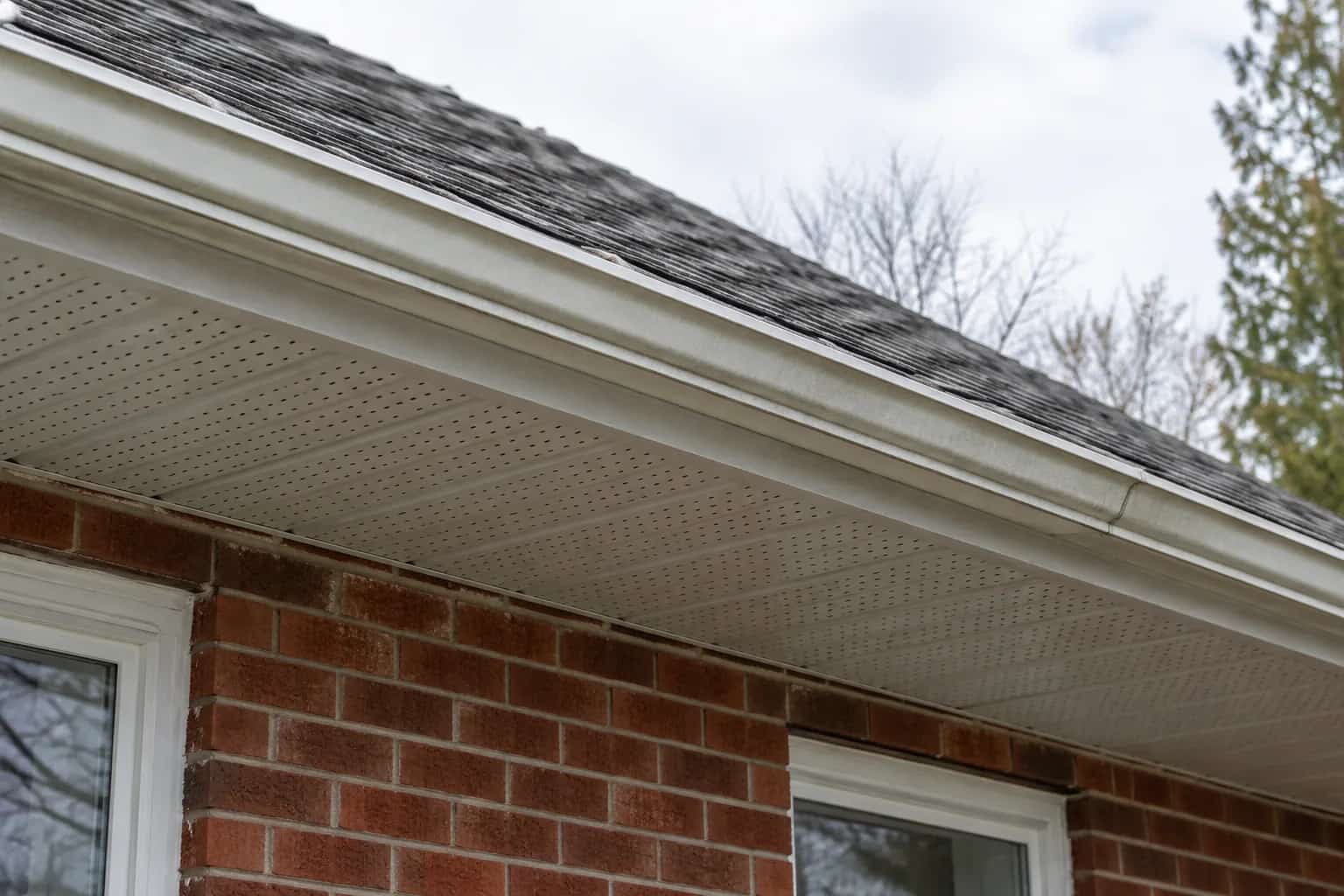 Perforated white soffit panels beneath an asphalt roof and eavestrough on a brick house